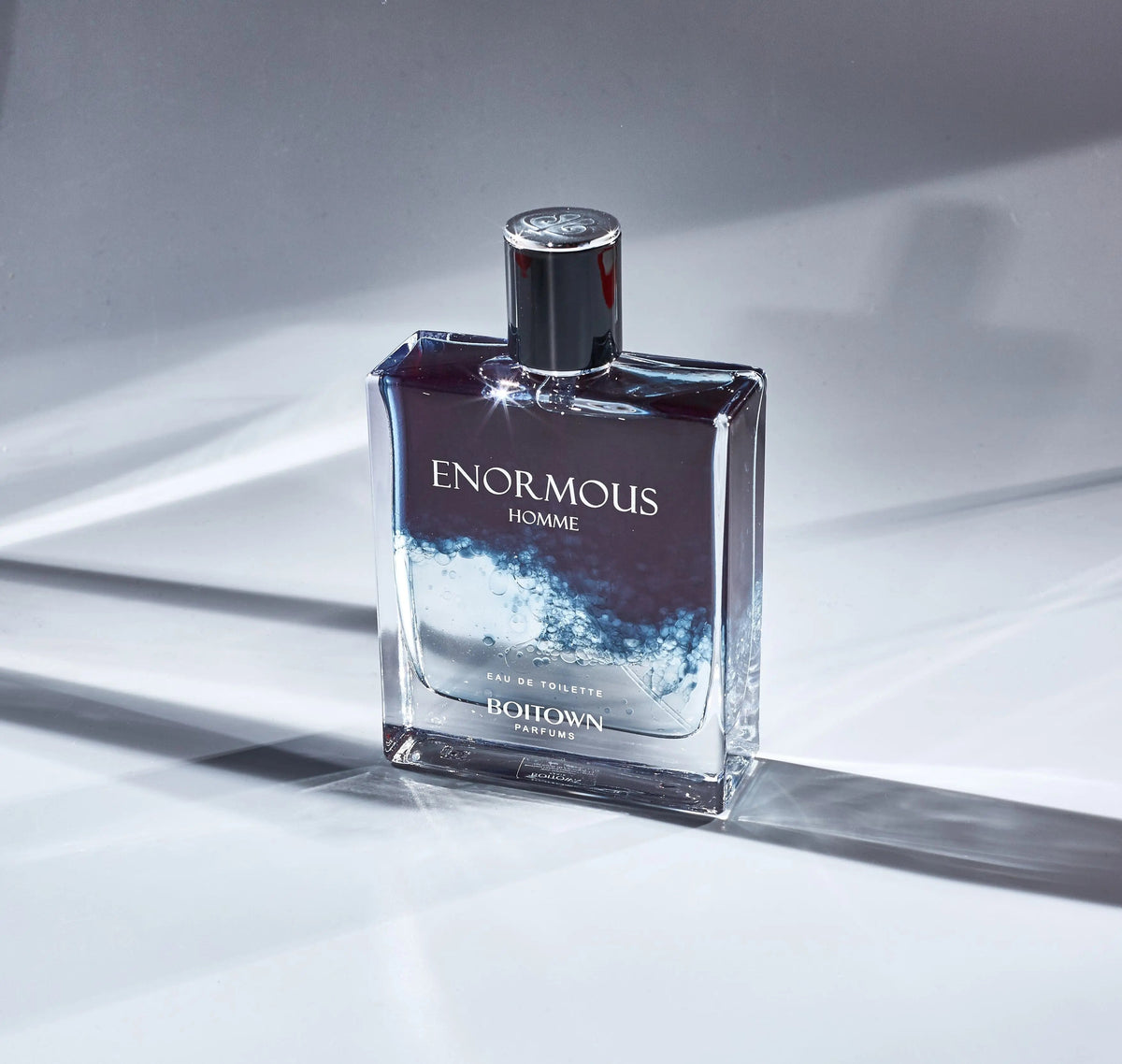 A rectangular glass bottle of Enormous Homme eau de toilette with a dark blue and clear liquid.