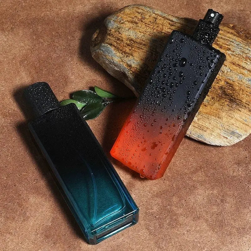 Two rectangular bottles of "Wholesale perfume scents long lasting fragrances men cologne fragrance," one blue-green and one red-orange with water droplets, rest on a brown surface beside wood and a green branch for an elegant display.