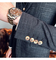 A man wears a dark grey striped three-piece suit with a luxury wristwatch on his wrist.