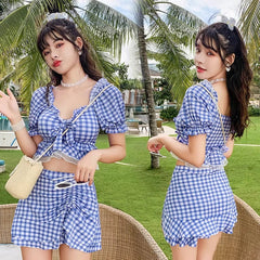 A woman wears a blue and white gingham two-piece swimsuit featuring a square-neck top with ruffled sleeves and a matching skirted bottom.