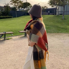 A woman wears a long, thick, multicolored plaid cashmere shawl with fringe in an outdoor setting.