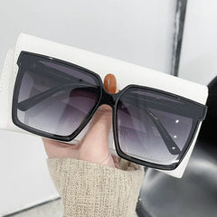 A hand holds a pair of oversized black square sunglasses with gradient lenses.