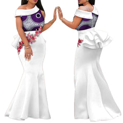 A woman models a white floor-length mermaid dress with a purple patterned bodice and floral applique.