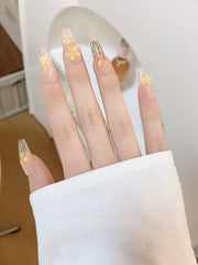 A hand displays long, clear press-on nails decorated with delicate yellow flowers and silver accents.