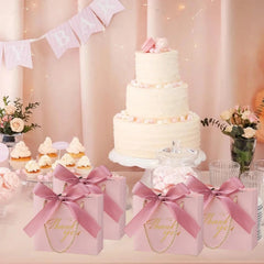 Four light pink mini gift bags with gold Thank You text and satin bows on a party table.