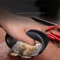 A hand uses a curved stainless steel garlic press to mince garlic on a wooden cutting board.