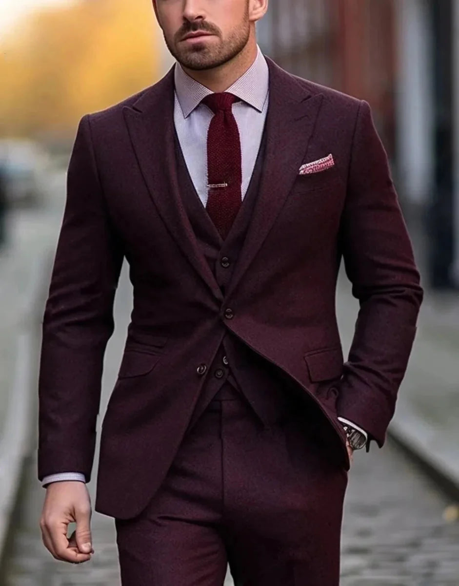 A man wears a burgundy three-piece suit with a matching tie and a light patterned dress shirt.