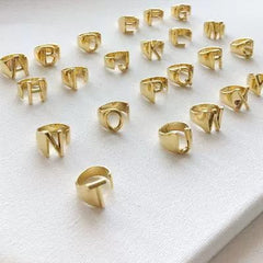 A collection of gold-plated adjustable rings featuring various uppercase letters arranged on a white surface.